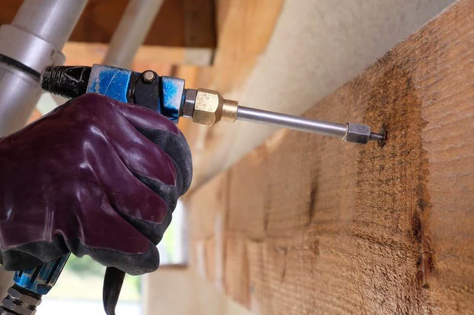 Artisan professionnel effectuant une inspection détaillée d'une charpente en bois à l'intérieur d'un comble ou d'une structure bâtie, équipé d'outils de diagnostic, examinant attentivement les poutres et les zones d'intersection pour détecter les dommages, les insectes xylophages ou les traces d'humidité. La lumière naturelle provient de fenêtres de toit, révélant la complexité de la structure en bois. L'artisan porte une tenue professionnelle appropriée et concentrée sur son diagnostic.